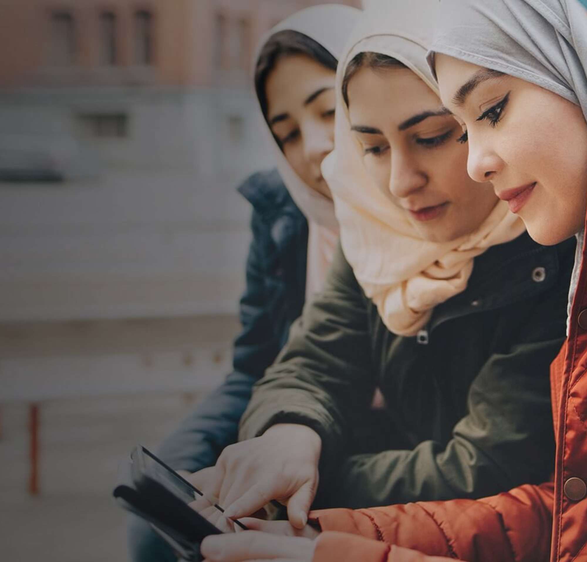 Four young women on a bench outside looking at a mobile device together.